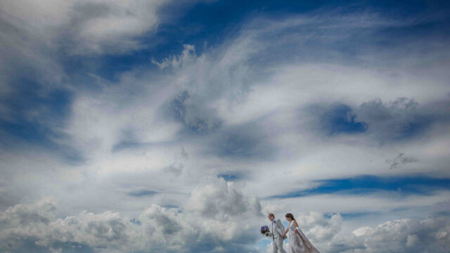 A couple in wedding attire walks on a rocky path against a vast, cloudy blue sky. The bride holds a bouquet.