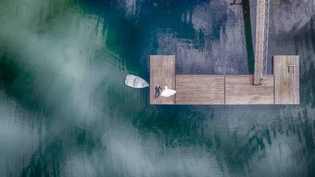 A couple in wedding attire lies on a wood dock next to a small rowboat, surrounded by calm, turquoise water with a ladder extending into the water from the dock.