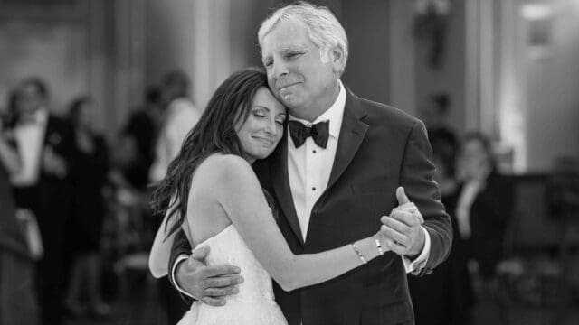 A bride and an older man in formal attire are embracing and dancing in a ballroom.