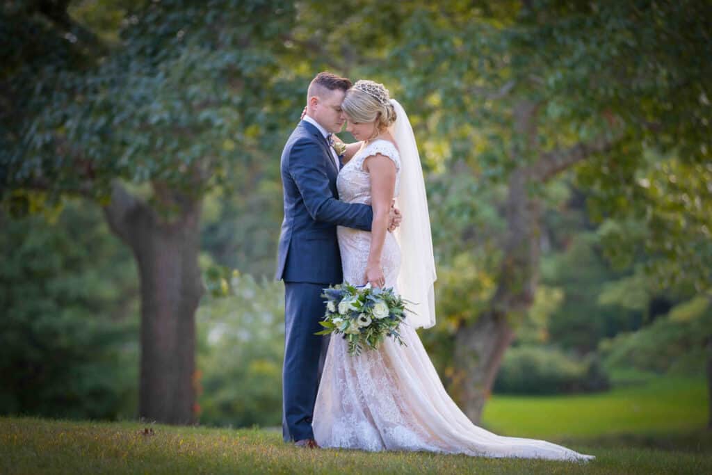 Bride and groom embrace outdoors, touching foreheads. The bride holds a green and white bouquet, with trees and grass visible in the background.