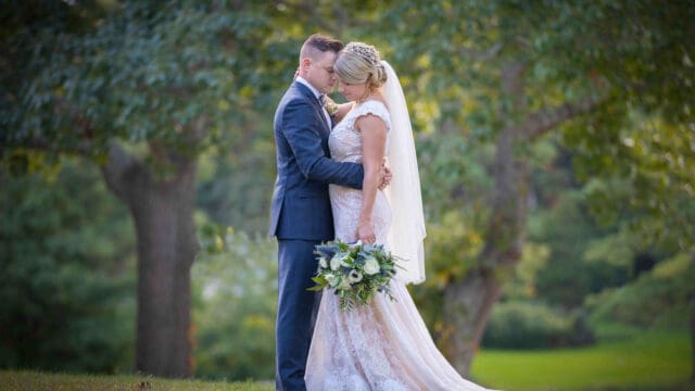 Bride and groom embrace outdoors, touching foreheads. The bride holds a green and white bouquet, with trees and grass visible in the background.