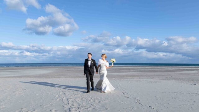 A bride and groom walk hand in hand on a sandy beach under a partly cloudy sky. The bride holds a bouquet of flowers.