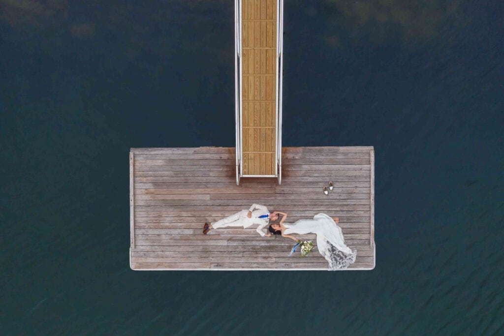 Aerial view of a bride and groom in white attire lying on a wooden dock protruding into a body of dark water.