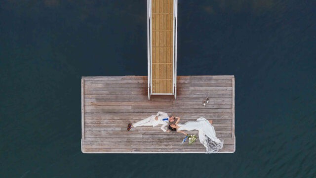 Aerial view of a bride and groom in white attire lying on a wooden dock protruding into a body of dark water.