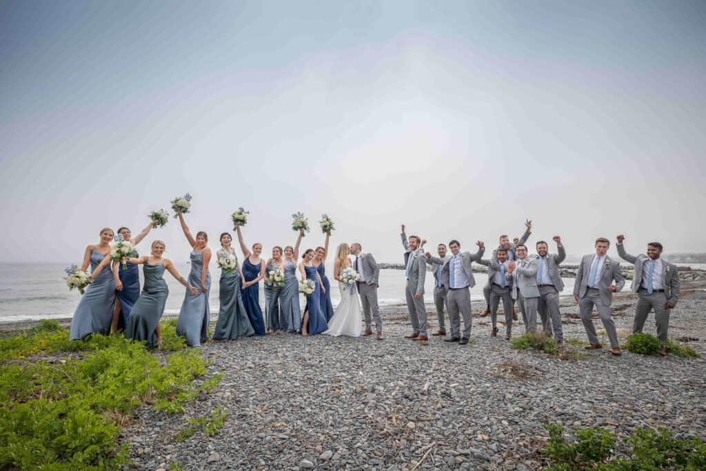 A large wedding party poses on a rocky beach, arms and bouquets raised. The group includes the bride, groom, bridesmaids, and groomsmen in coordinating attire.