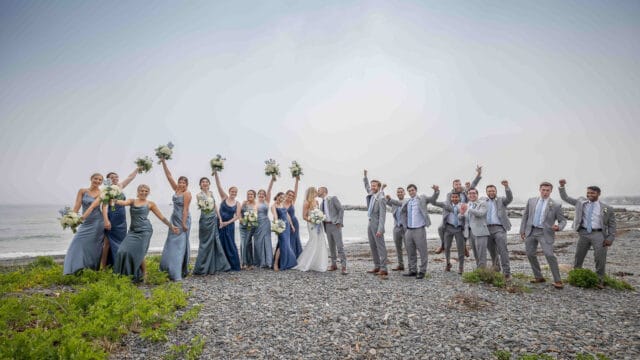 A large wedding party poses on a rocky beach, arms and bouquets raised. The group includes the bride, groom, bridesmaids, and groomsmen in coordinating attire.