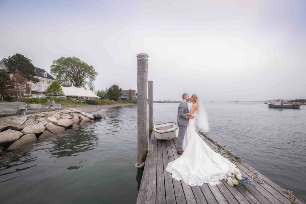 A bride and groom stand on a wood dock by the water, embracing and kissing. A small boat is tied to the dock, with buildings and trees visible in the background.