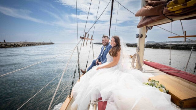 A bride and groom sit together on the deck of a sailboat, dressed in wedding attire, with the sea and cloudy sky in the background.