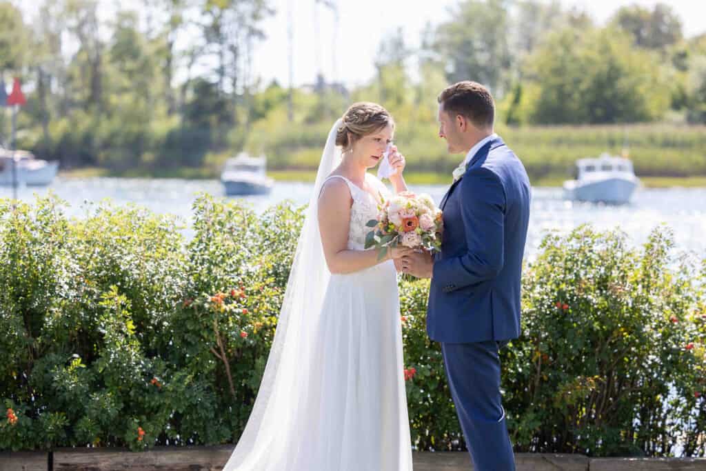 Bride and groom stand by the water, smiling as the bride wipes a tear. They hold a bouquet, with boats visible in the background.