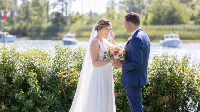 Bride and groom stand by the water, smiling as the bride wipes a tear. They hold a bouquet, with boats visible in the background.