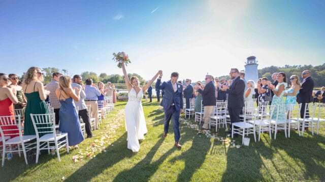 A bride and groom hold hands and walk down an outdoor aisle, surrounded by standing guests and white chairs, on a sunny day. The bride holds a bouquet in the air.