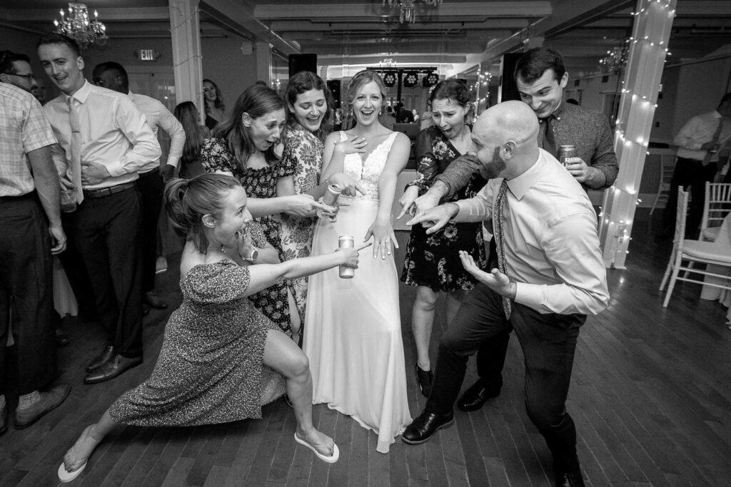 A group surrounds the bride, some pointing in surprise while others smile and gesture. The scene is in a reception hall, with a celebratory mood.