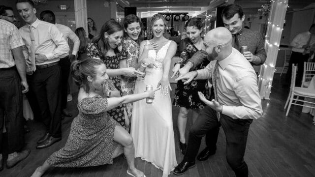 A group surrounds the bride, some pointing in surprise while others smile and gesture. The scene is in a reception hall, with a celebratory mood.