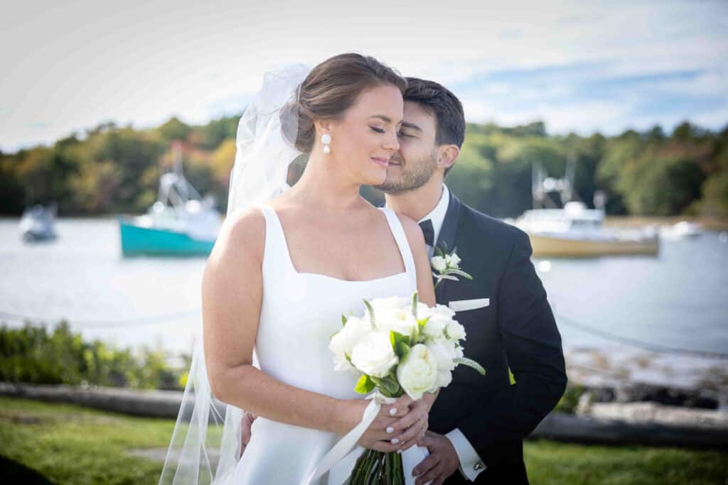 Bride and groom stand by a lakeside, the groom kissing her cheek as she holds a white bouquet. Boats and trees are visible in the background.