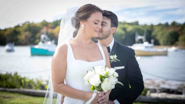 Bride and groom stand by a lakeside, the groom kissing her cheek as she holds a white bouquet. Boats and trees are visible in the background.