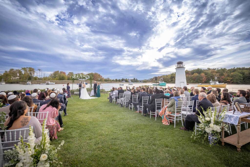 Wedding ceremony by a river with a lighthouse in the background. Guests sit on white chairs on grass, facing the couple and officiant under an archway.