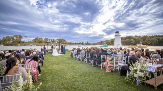 Wedding ceremony by a river with a lighthouse in the background. Guests sit on white chairs on grass, facing the couple and officiant under an archway.