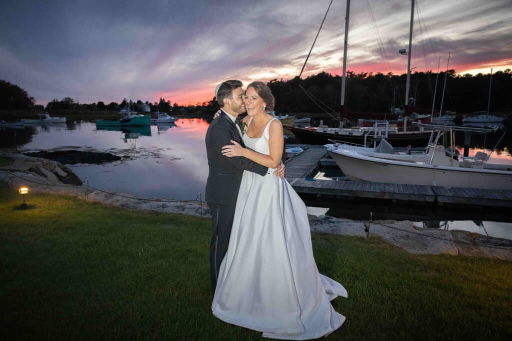 A couple, dressed in formal attire, embraces near a marina at sunset with docked boats and a picturesque sky in the background.