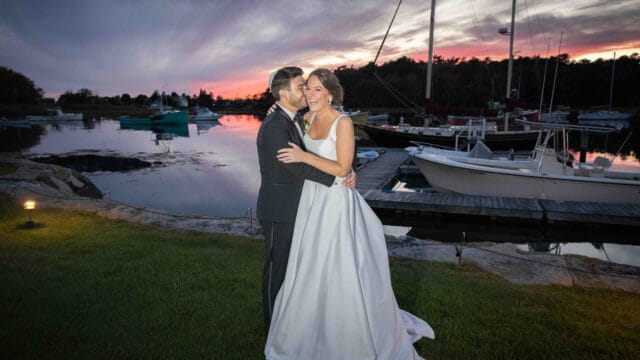 A couple, dressed in formal attire, embraces near a marina at sunset with docked boats and a picturesque sky in the background.