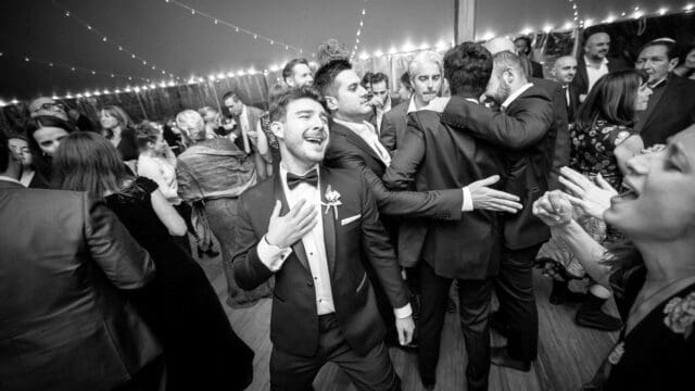 A man in a tux joyfully dances in the foreground at an indoor event with string lights, while others socialize in the background.