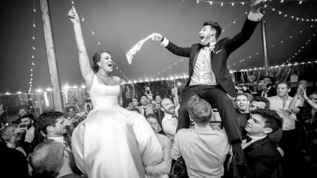 The bride and groom are lifted up by guests at their wedding reception, celebrating joyfully with string lights and cheering guests in the background.