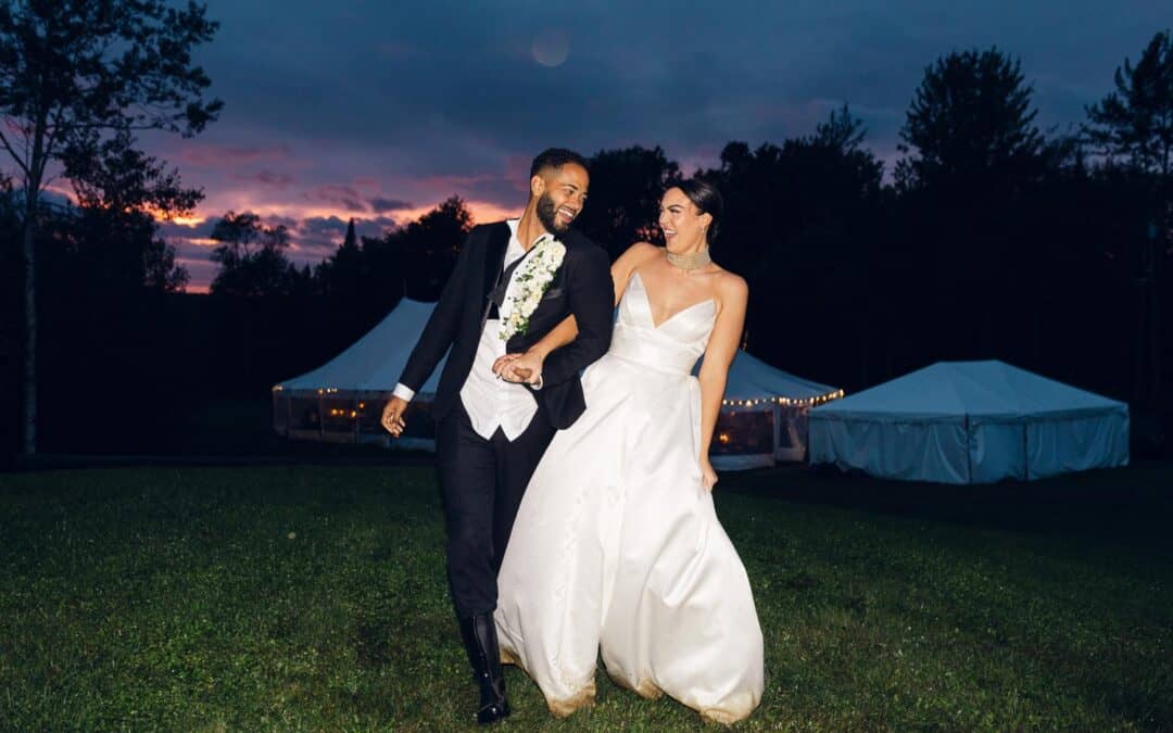 A couple, dressed in formal wedding attire, smiling and holding hands while walking on a grassy area at dusk with tents and a dark sky in the background.