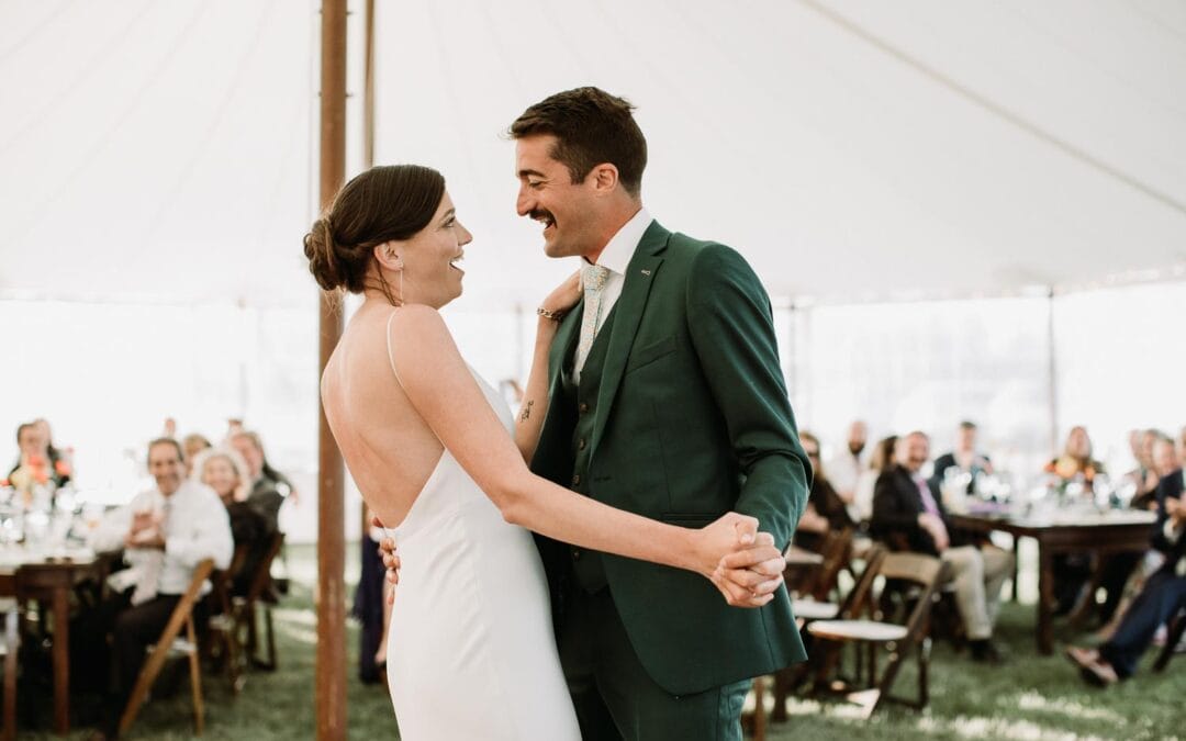 A couple, dressed in wedding attire, dances together under a tent with guests seated at tables in the background.