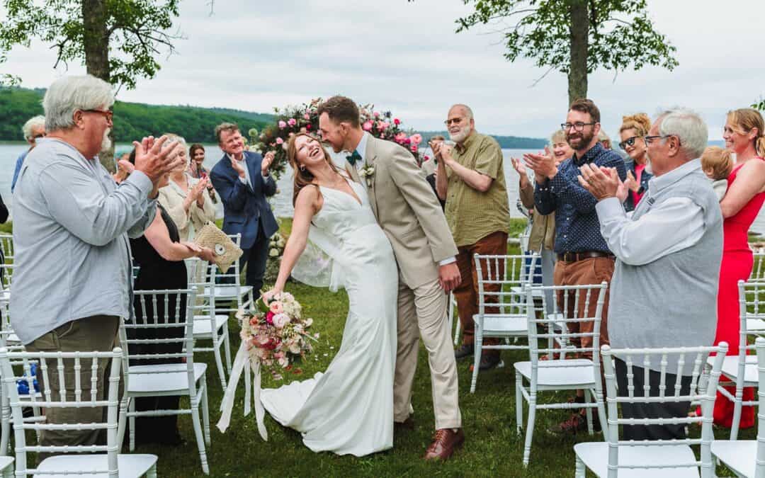 A bride and groom share a kiss while walking down the aisle, surrounded by clapping guests at an outdoor wedding ceremony.