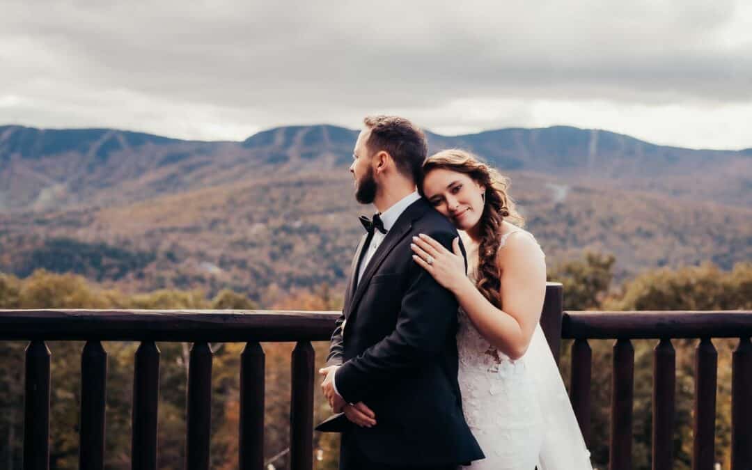 A bride in a white wedding dress leans her head on the groom's shoulder, as he stands facing slightly away, against a mountainous backdrop with cloudy skies.