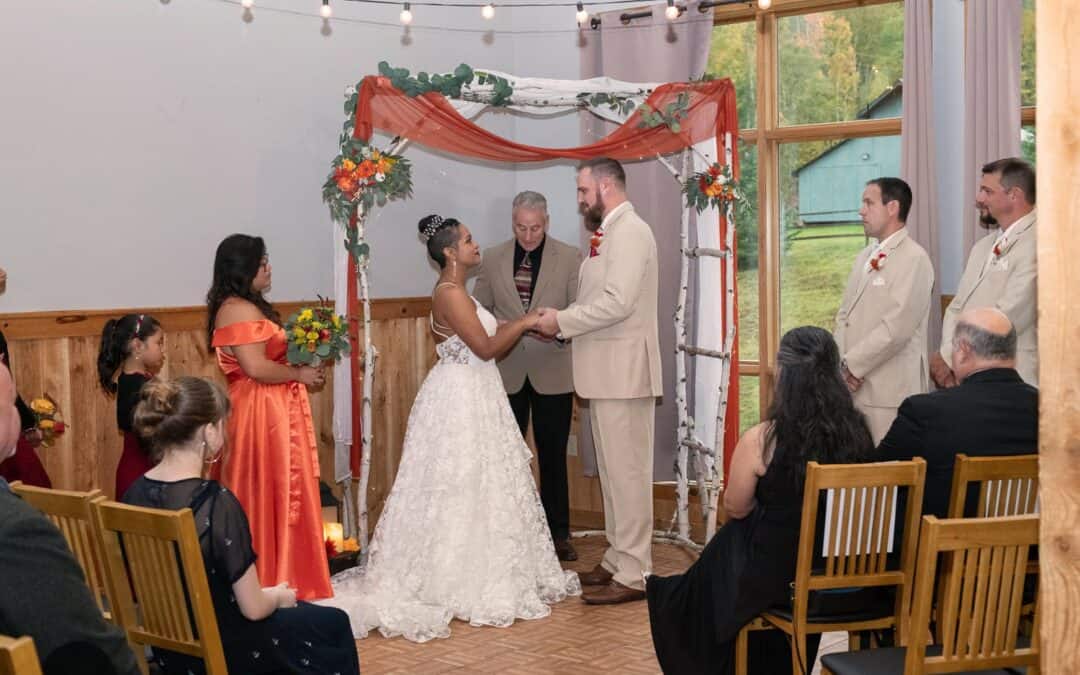 Bride and groom stand under a decorated arch exchanging vows, holding hands. Guests, bridesmaids, and groomsmen observe in an indoor setting with a large window.