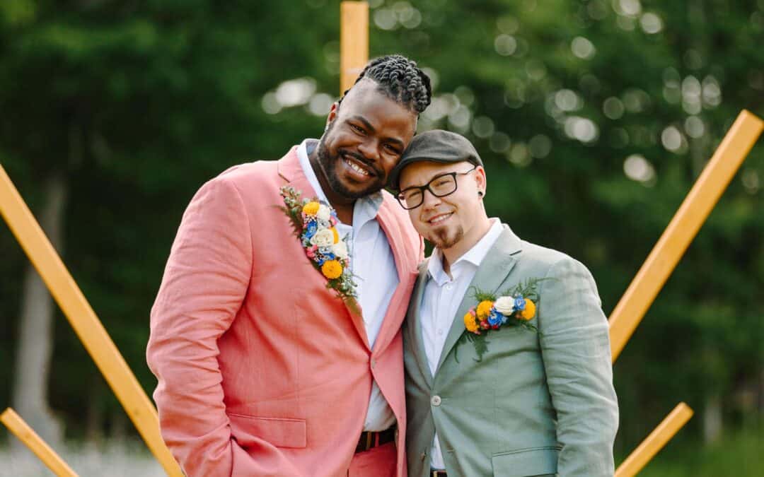 Two people outdoors in pastel suits with matching floral boutonnieres; one in coral, the other in light green. A geometric wooden structure stands behind them.