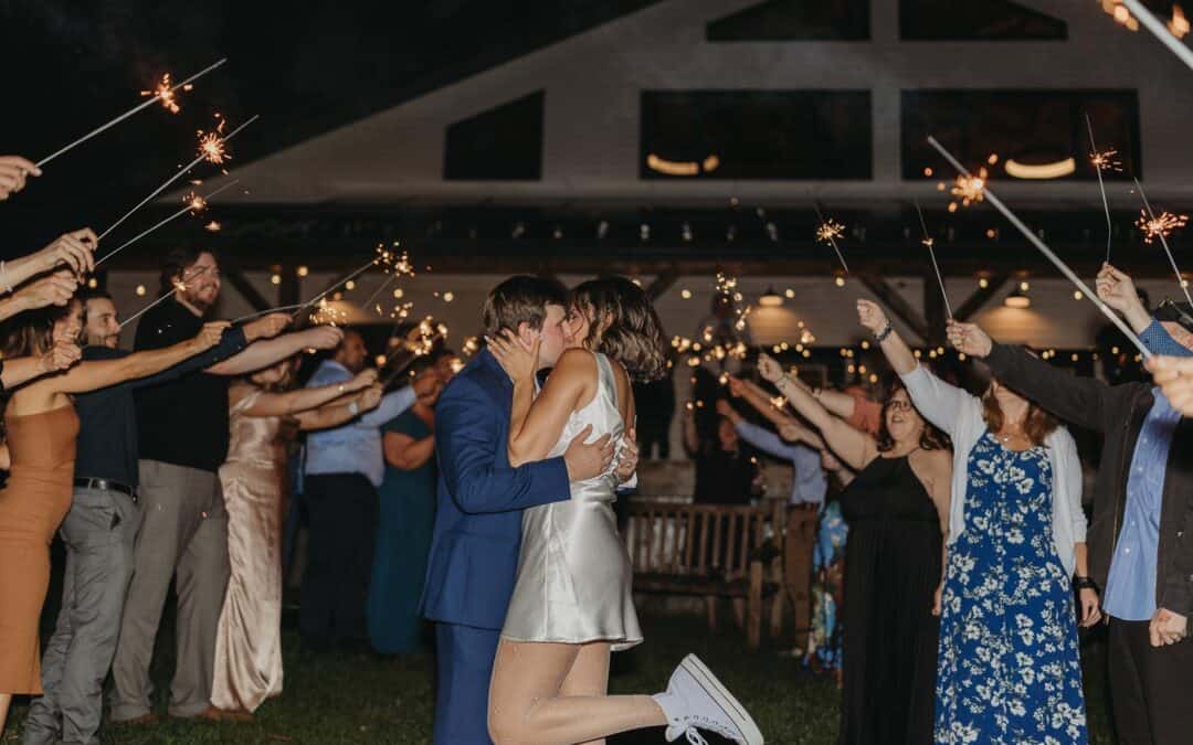 A couple kisses while guests hold sparklers in front of a house during an outdoor evening celebration.