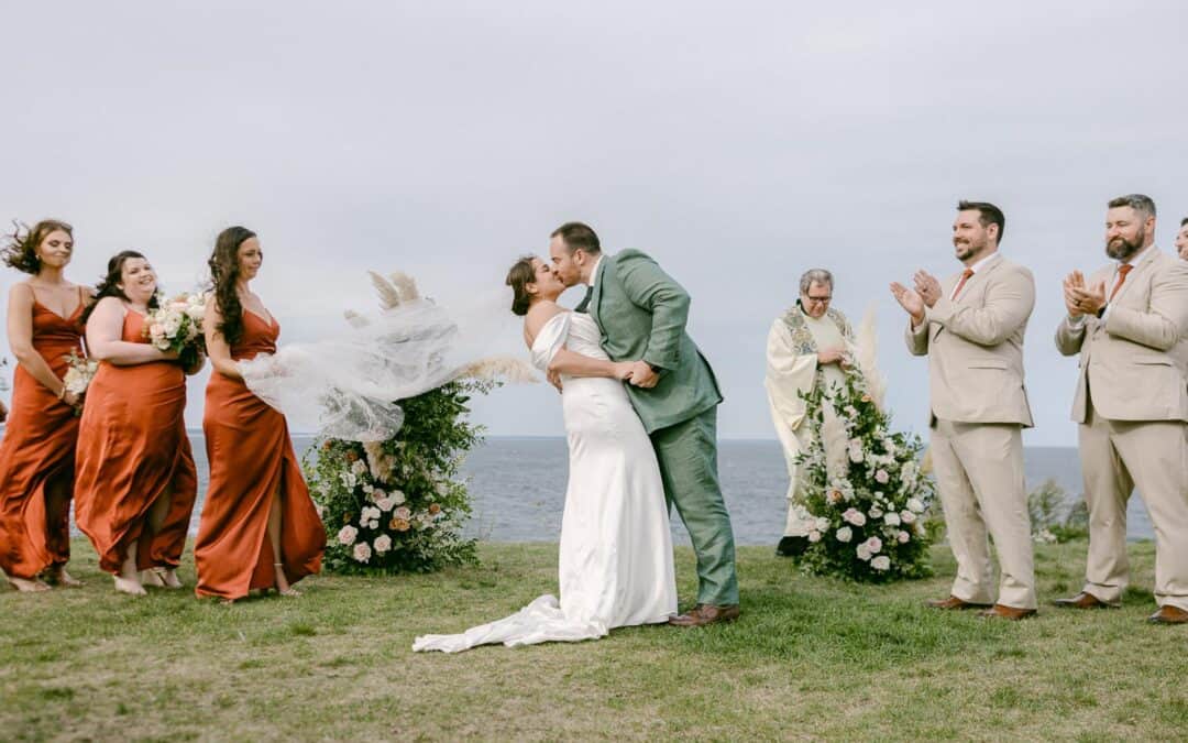 A couple shares a kiss during their outdoor wedding with bridesmaids in orange dresses and groomsmen in beige suits nearby, alongside a preacher.